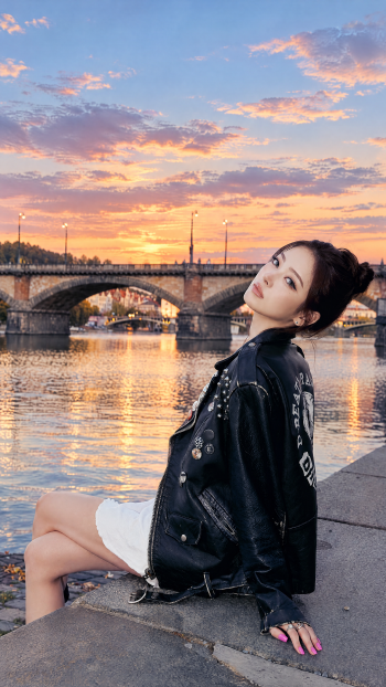MEOVV “GO HARD” woman portrait iPhone wallpaper, seated by a riverside bridge at sunset in a black leather jacket.