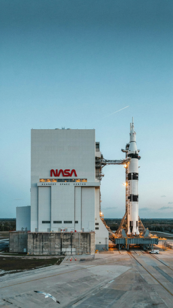 NASA rocket on the launchpad beside the Vehicle Assembly Building under a clear blue sky, framed as an iPhone space wallpaper.
