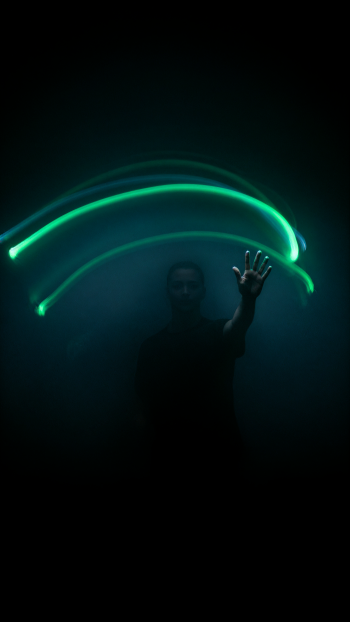 Silhouette waving beneath vivid green light trails in a dark, atmospheric light painting phone wallpaper.