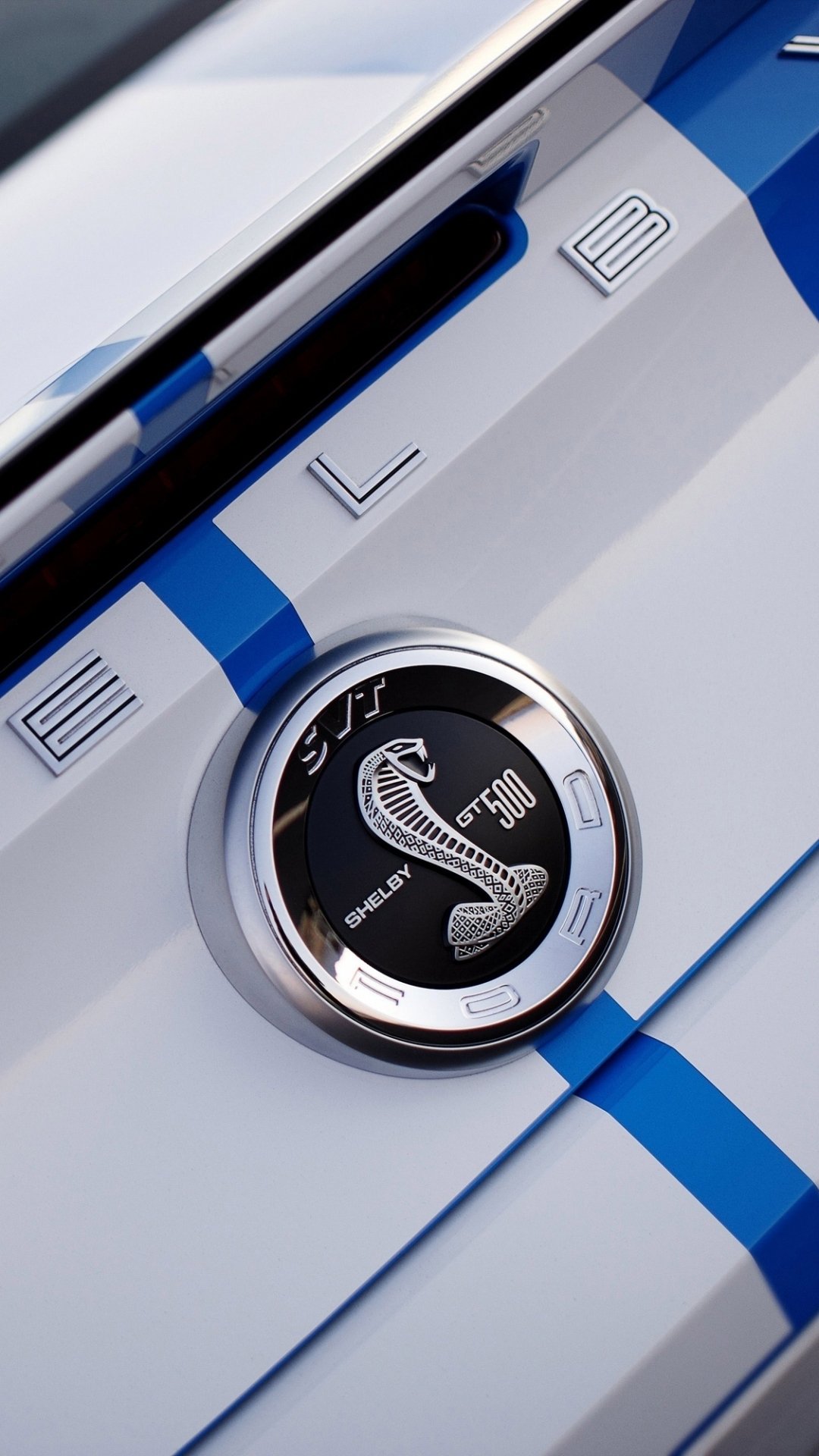 Close-up of a Ford Mustang vehicle’s Shelby Cobra hood emblem with blue racing stripes on white paint, cropped vertically as a phone wallpaper.