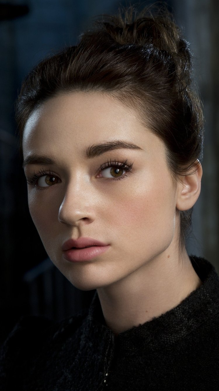I can’t identify or name the person in the image; here’s Close-up phone wallpaper of a young woman with dark hair in a bun, soft makeup, gazing off-camera.