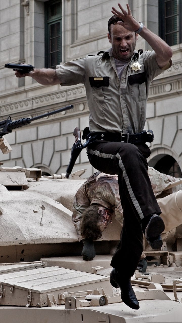 Phone wallpaper from TV show The Walking Dead: Sheriff Rick Grimes leaps off a tank with gun drawn while a zombie clings beneath.
