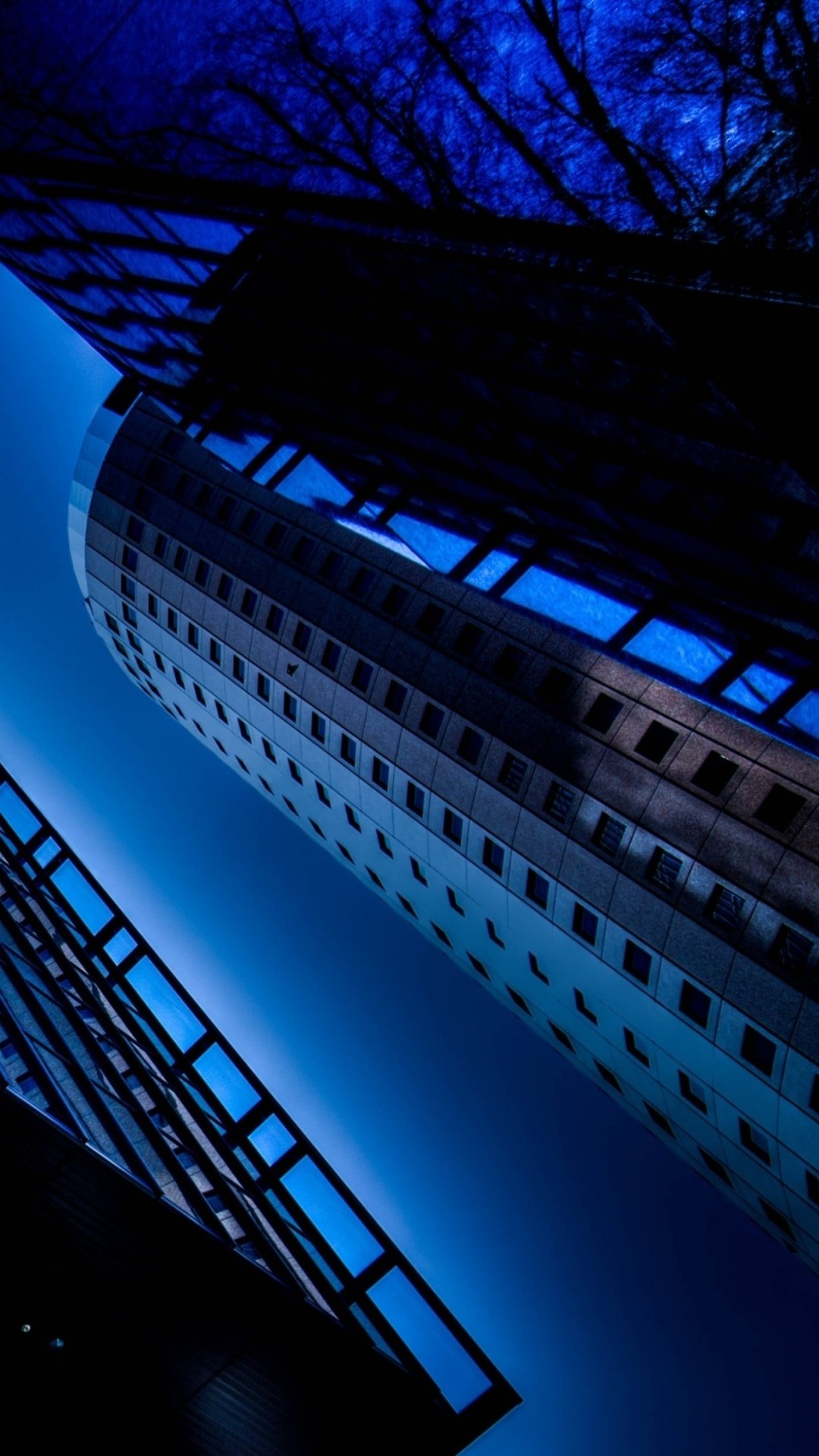 Vertical phone wallpaper of a Tokyo, Japan skyscraper — blue-toned, towering man-made architecture framed by angular buildings and bare tree branches against a deep night sky.