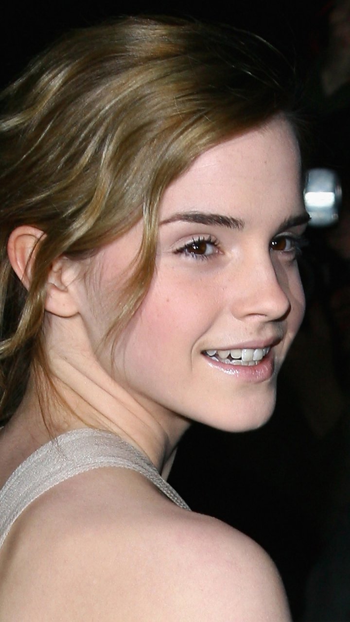 Phone wallpaper close-up of a young woman smiling over her shoulder, light brown hair swept back, wearing a sleeveless top, dark background.