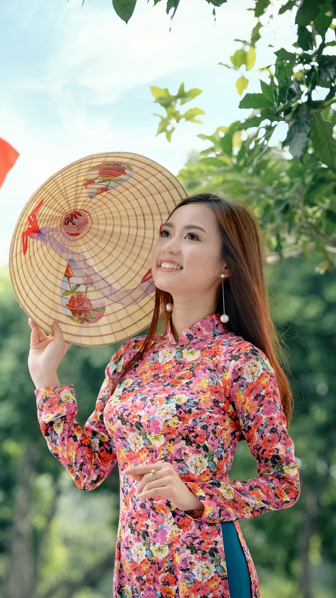 Smiling Asian woman with brown eyes and earrings wears a floral traditional costume and holds a decorated hat, set against a bright outdoor background.