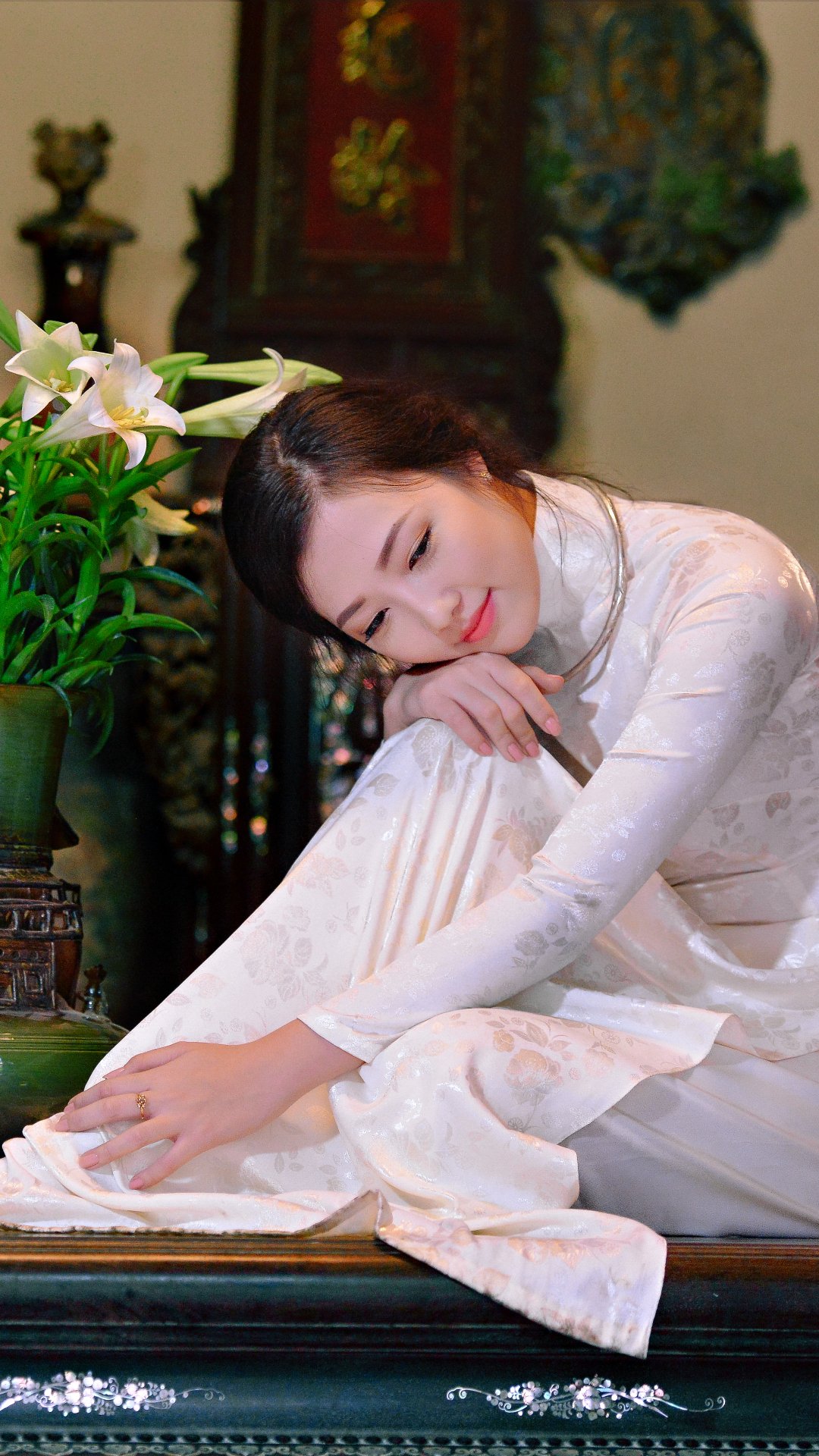 Asian Vietnamese model in a white Ao Dai gently touching lilies, captured in a serene moment with a warm, traditional backdrop, formatted for phone wallpapers.
