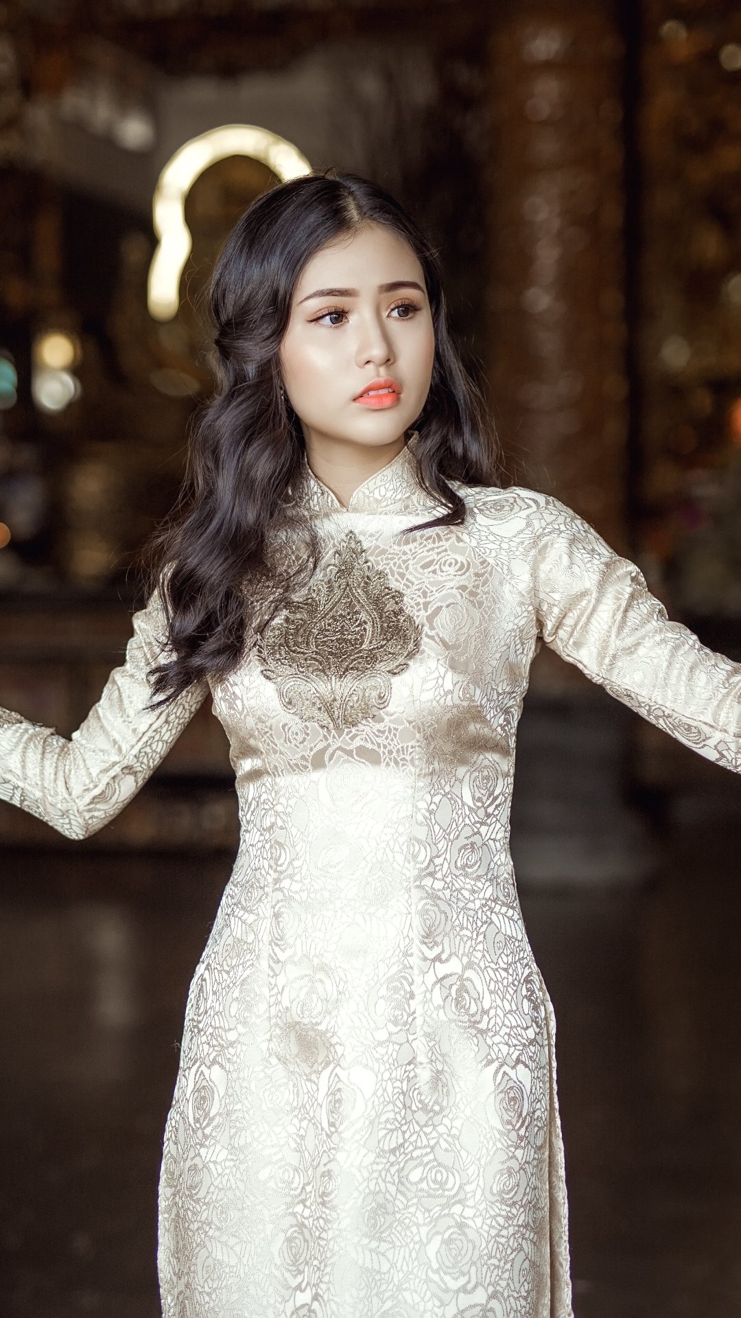 Phone wallpaper: portrait of a woman model in an ornate white dress with dark wavy hair, poised expression against a softly lit, decorative background.