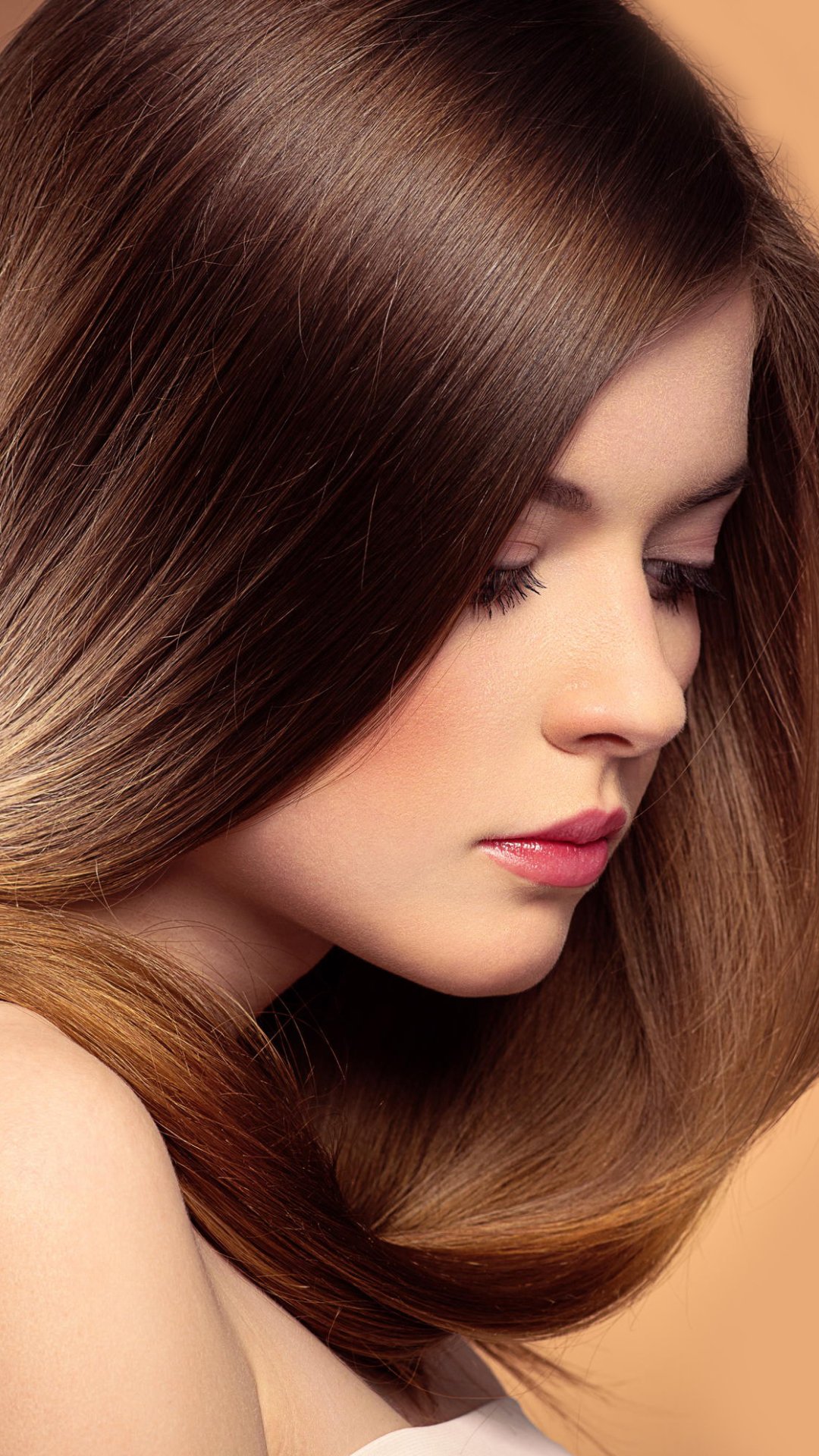 Phone wallpaper: close-up profile of a brunette woman's face, smooth hair framing a contemplative, serene mood in warm, soft tones.