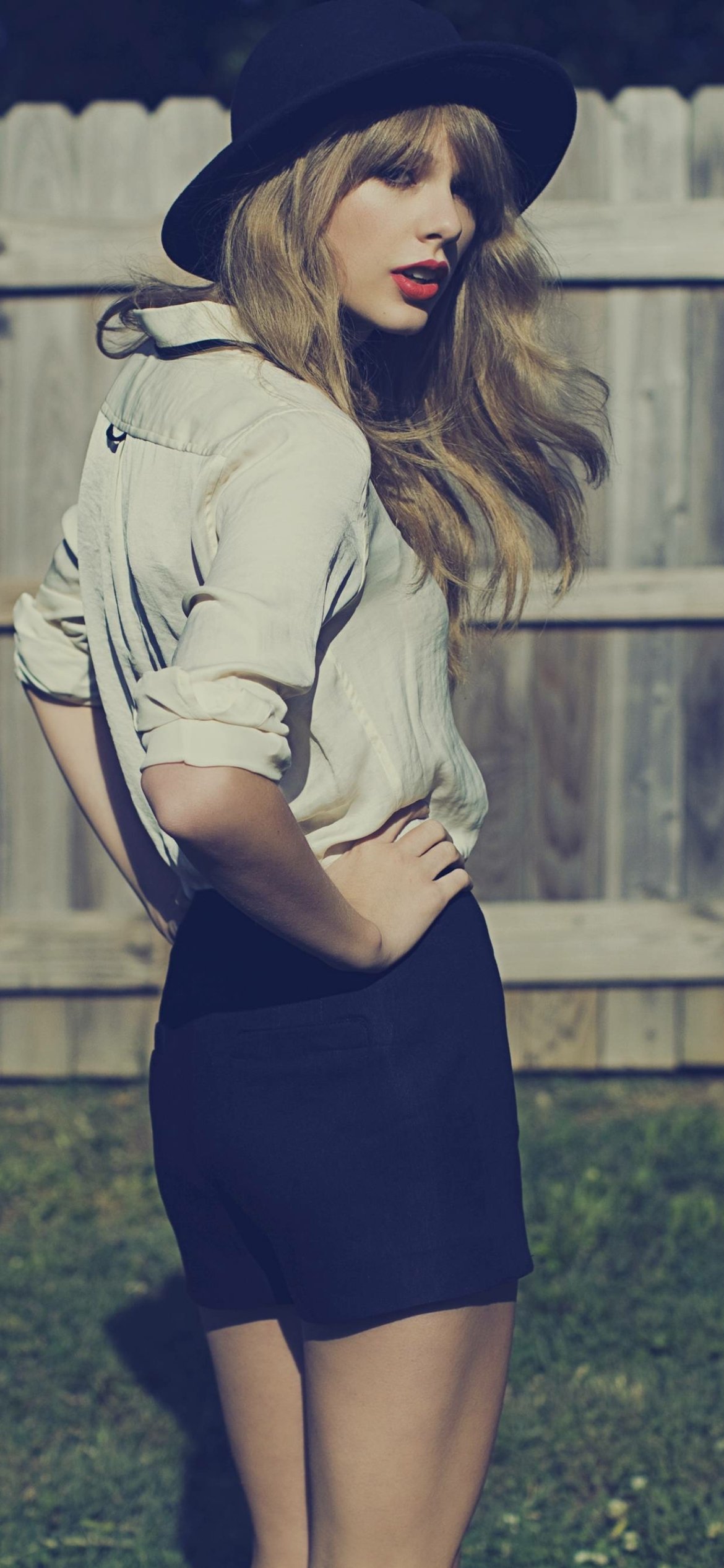 Phone wallpaper featuring Taylor Swift in a hat and stylish outfit, with a blurred wooden fence background, capturing her music star vibe for iPhones and Android devices.