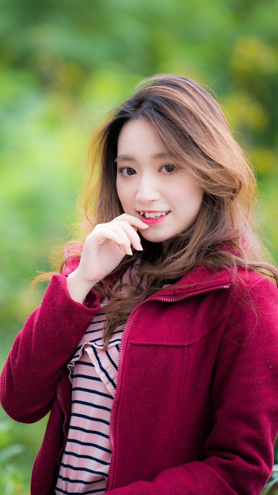 Phone wallpaper portrait of a smiling brunette model woman, shallow depth of field with soft green bokeh, hand near her chin, wearing a maroon coat over a striped top.
