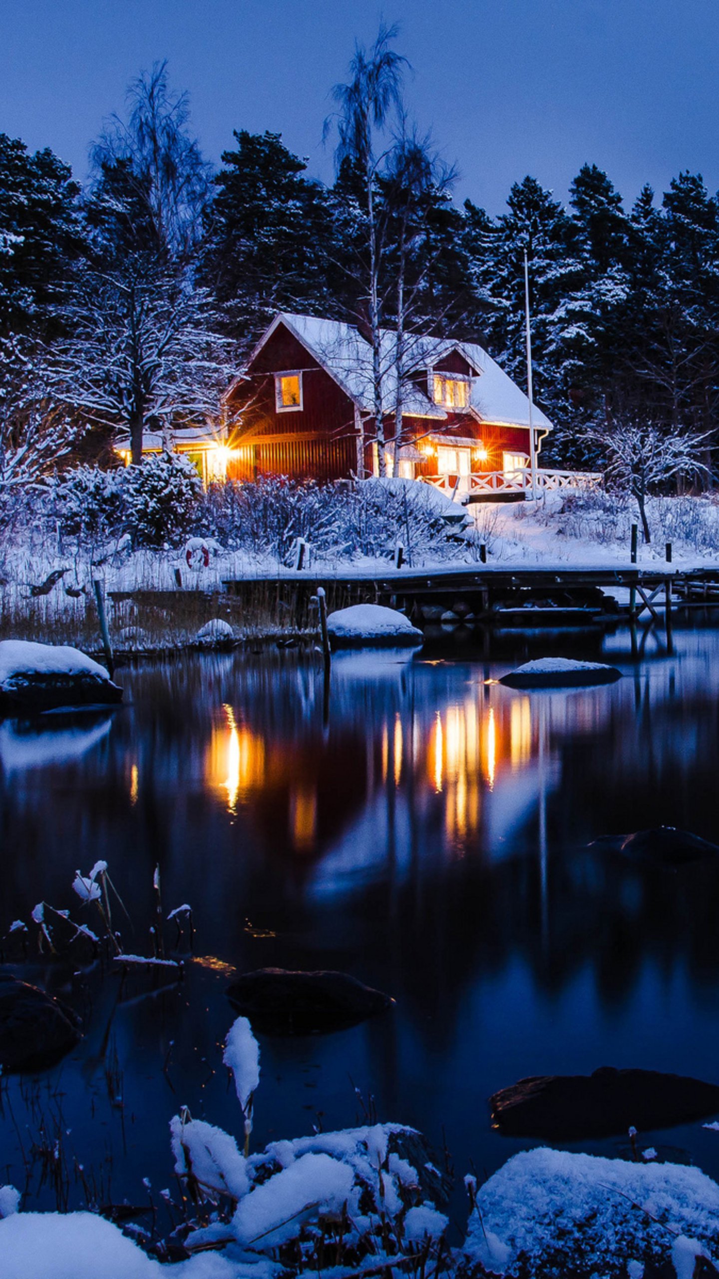 Phone wallpaper photograph of a snow-covered winter night: a warmly lit house by a calm lake, trees dusted with snow and their lights reflecting on the dark water.