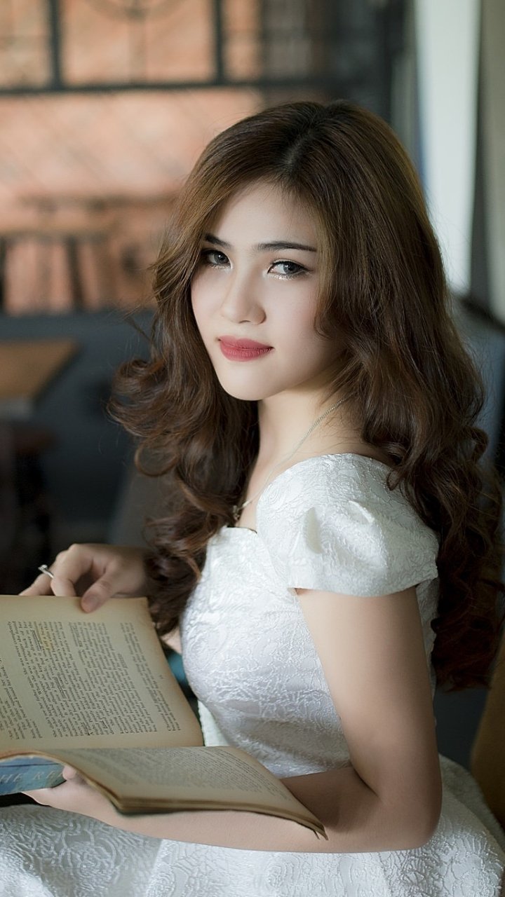 Portrait of an Asian brunette woman with brown eyes in a white dress, reading a book, designed as a phone wallpaper for iPhone and Android.