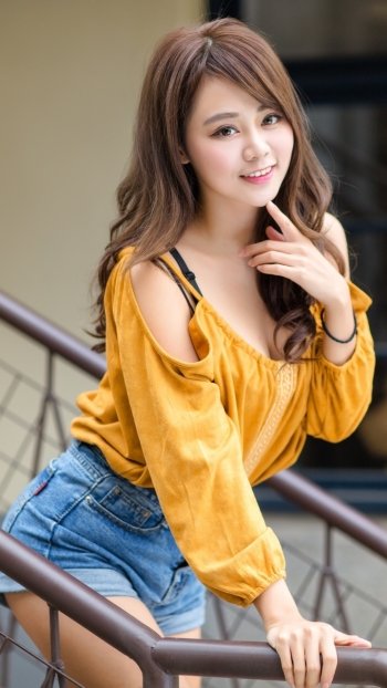 Phone wallpaper of a smiling brunette woman model in a yellow off-shoulder top and denim shorts, posing on a railing.