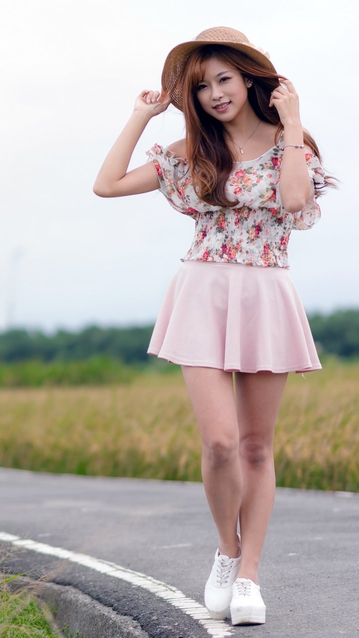 Brunette Asian woman in a floral top, pink skirt, and hat standing on a road with a blurred natural background, captured with depth of field.