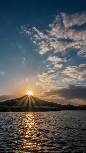 Vertical phone wallpaper of nature: sun and sunbeams rising over a mountain above a lake, sunlight reflecting on water under a vast sky with scattered clouds.