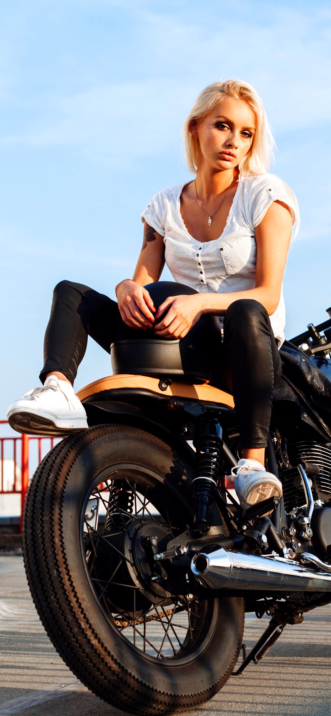 Phone wallpaper: blonde woman in a white top and black pants sitting on a café‑style motorcycle under a blue sky — Girls & Motorcycles.