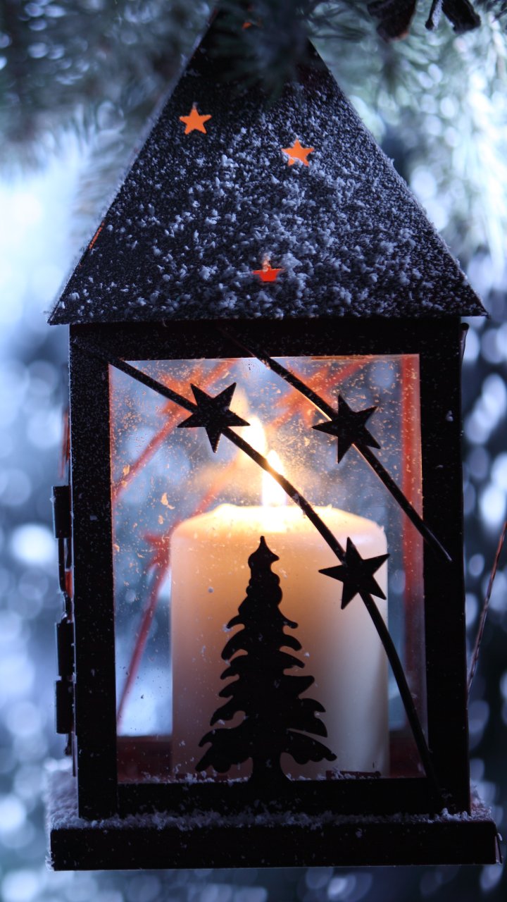 Warm candlelight glows inside a snow-dusted lantern with star cutouts and an evergreen silhouette — festive Christmas holiday phone wallpaper.