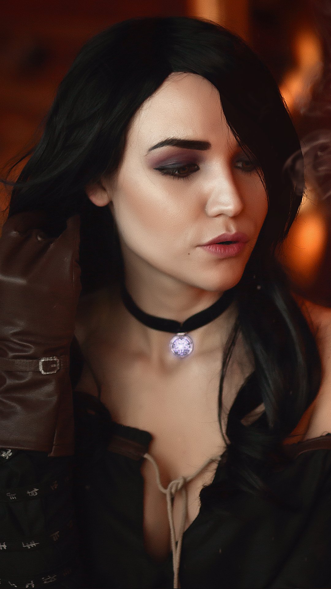 Phone wallpaper of a woman cosplaying Yennefer of Vengerberg (The Witcher 3: Wild Hunt): long black hair, black choker collar, dark outfit and gloves, looking down in warm light.