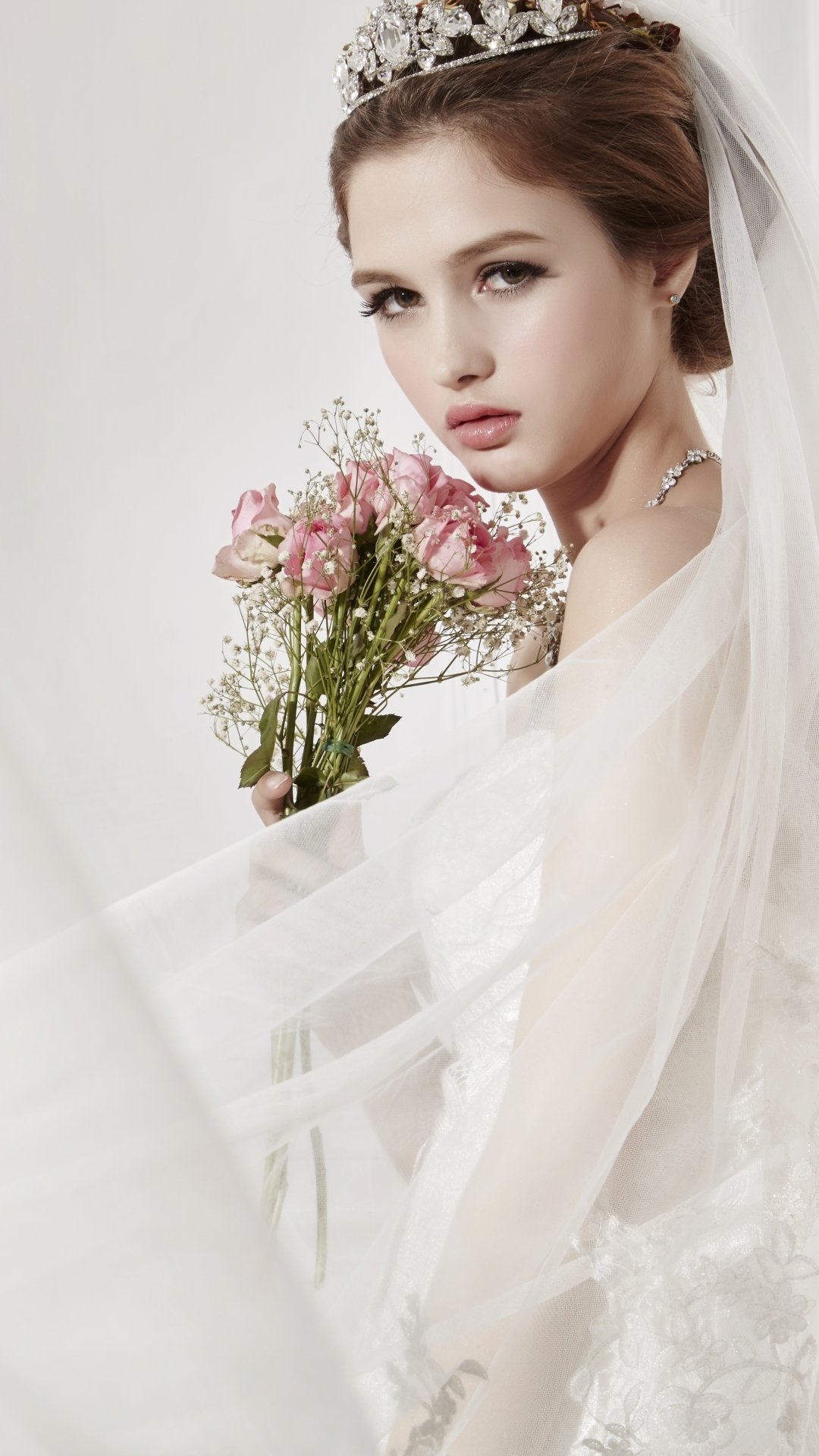 Bride in a wedding dress and tiara, brown-eyed woman holding pink flowers, soft veil framing her face — vertical phone wallpaper.