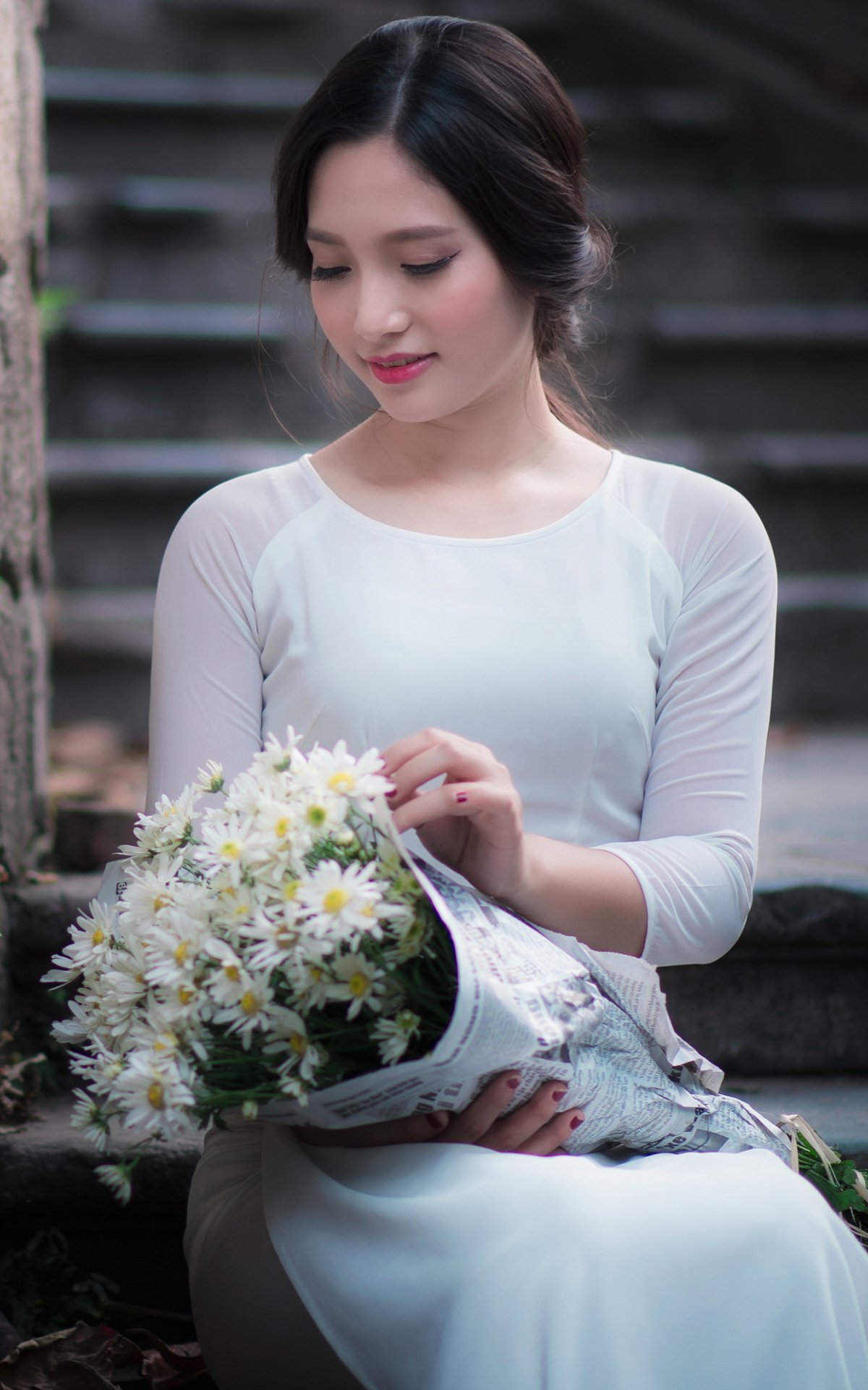 Asian woman with black hair in a white dress holding a bouquet of daisies, sitting thoughtfully on outdoor steps, captured in a serene mood for phone wallpaper.