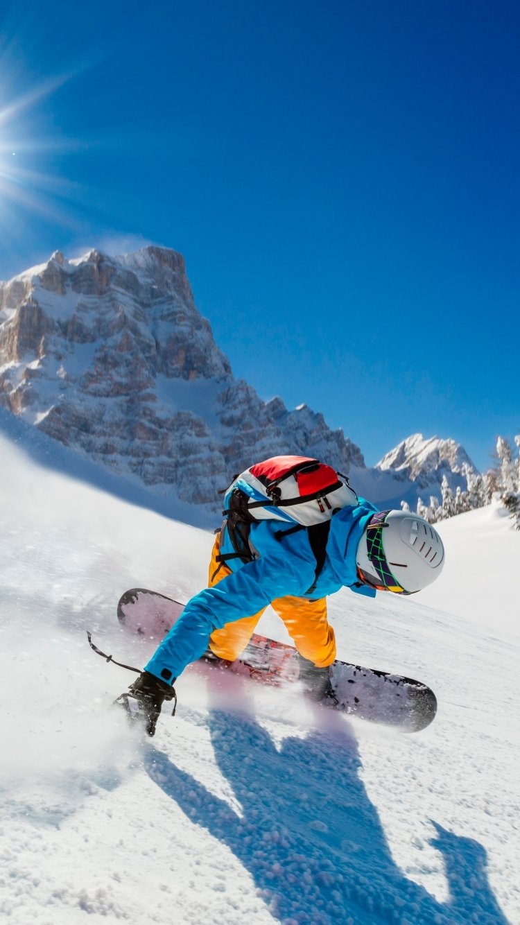 Phone wallpaper of a snowboarder carving through deep snow on a snowy mountain—winter sports scene under a bright blue sky with the sunburst over rugged peaks.
