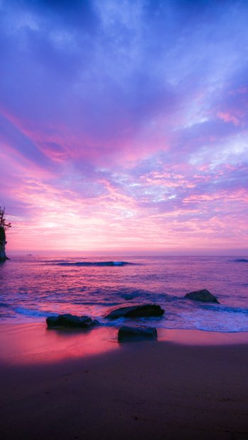 Vertical phone wallpaper of a tranquil beach at sunset: pink-purple sky over ocean and sea, soft waves against rocks on the sandy shore meeting the distant horizon.