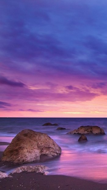Phone wallpaper showing a tranquil beach with rocks, a vivid purple-pink sky meeting the horizon, and calm ocean washing the earthy shoreline.