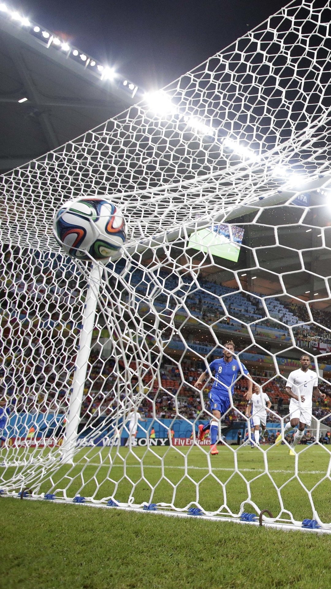 Phone wallpaper showing a soccer ball hitting the net in a stadium, players and crowd blurred beneath bright floodlights — dynamic sports action.