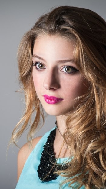 Phone wallpaper of a long-haired woman model with bright pink lipstick and a black statement necklace, gazing at the camera against a soft gray background.