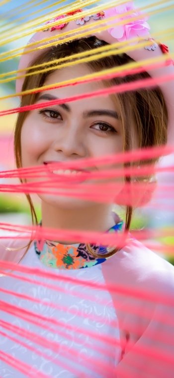 Phone wallpaper portrait of a smiling Asian brunette woman model with brown eyes, soft blur and colorful threads across the frame, compatible with iPhone and Android screens.