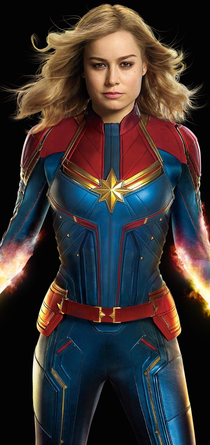 Phone wallpaper of Captain Marvel from the movie: a stern, windswept blonde superhero in blue-red-gold armor with glowing fists against a dark background.