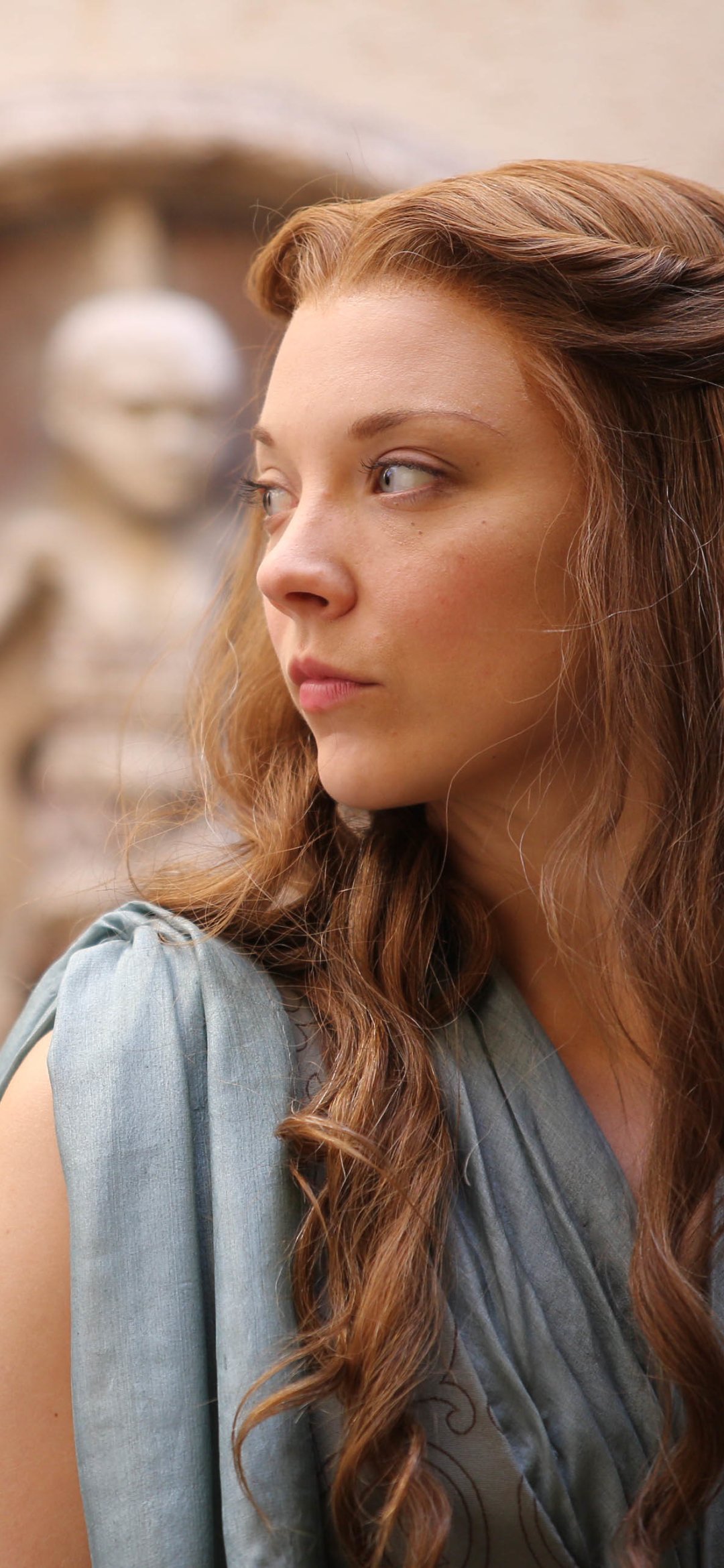 Margaery Tyrell from Game of Thrones as a phone wallpaper: side-profile portrait with long curled chestnut hair, pale blue gown, and a blurred stone courtyard background.