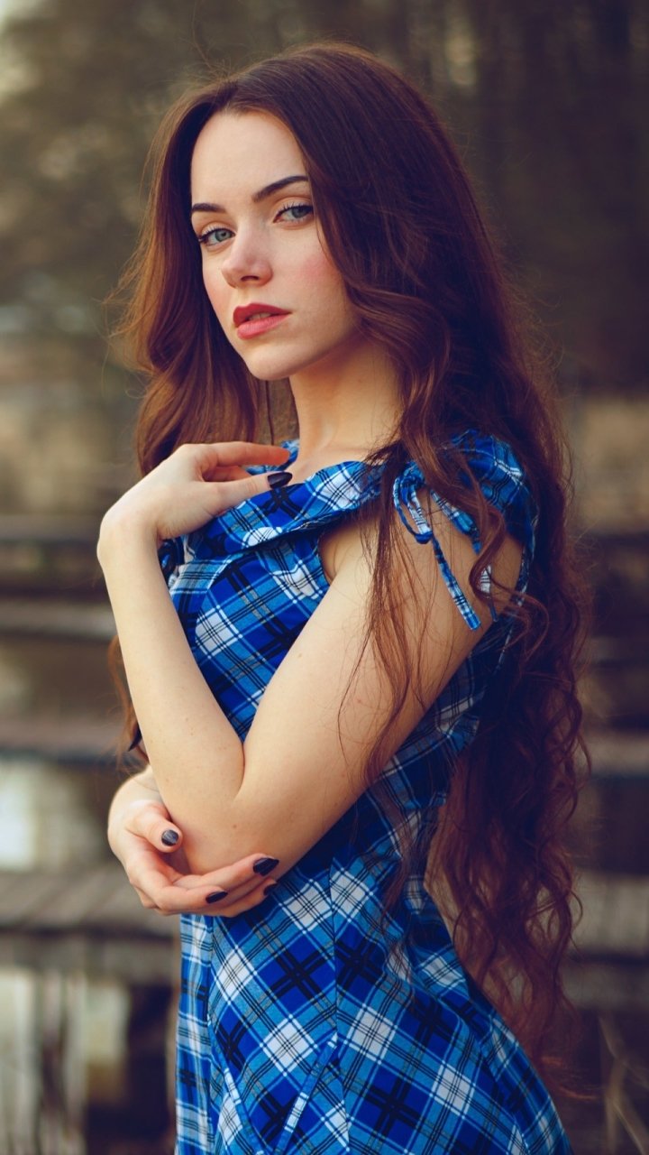 Phone wallpaper of a redhead woman model in a blue plaid dress, long wavy hair flowing over her shoulders, posing outdoors against a soft, blurred background.