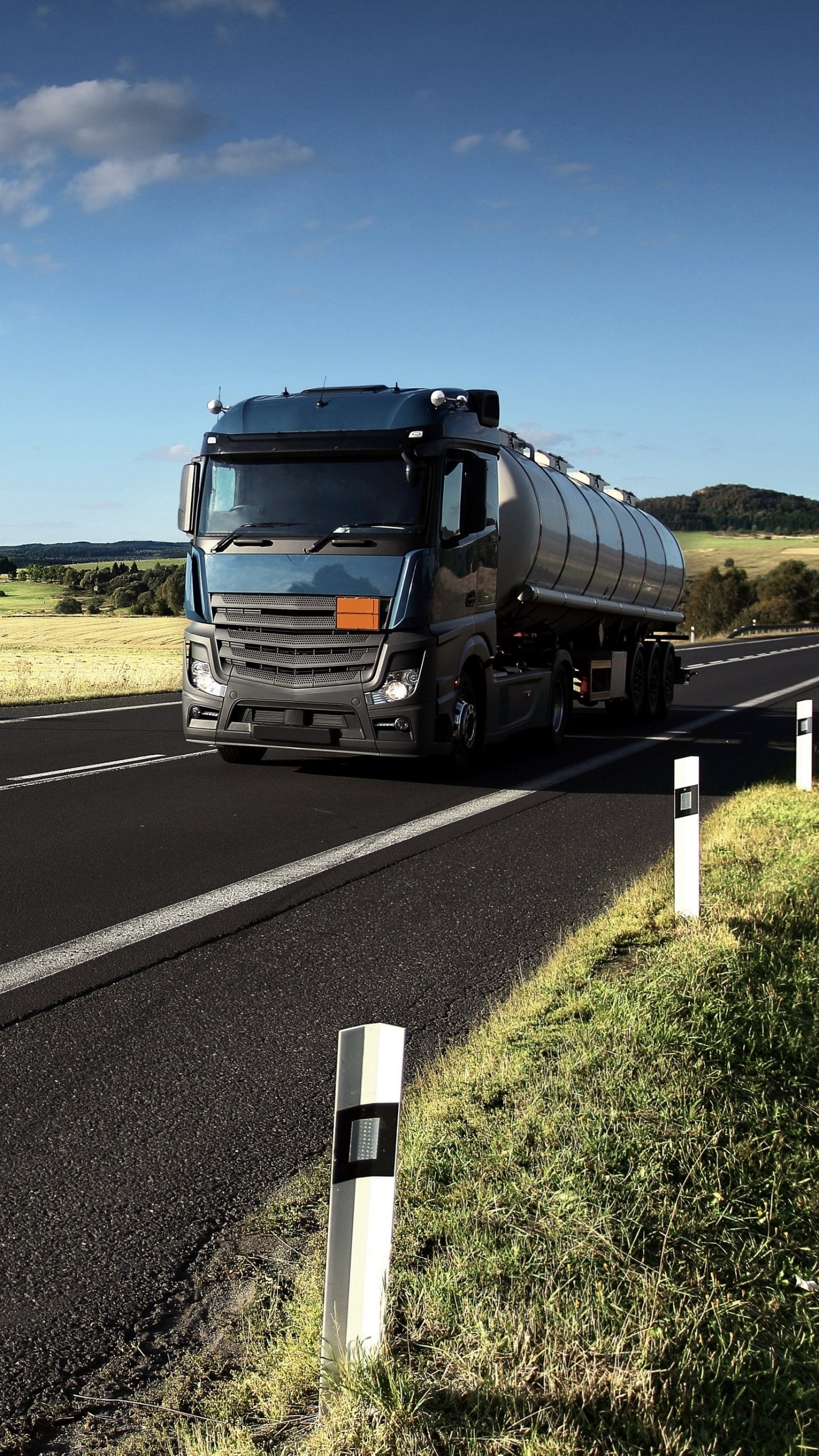 Phone wallpaper showing a truck tanker vehicle on a sunlit country highway under a blue sky, with roadside markers and rolling green fields.