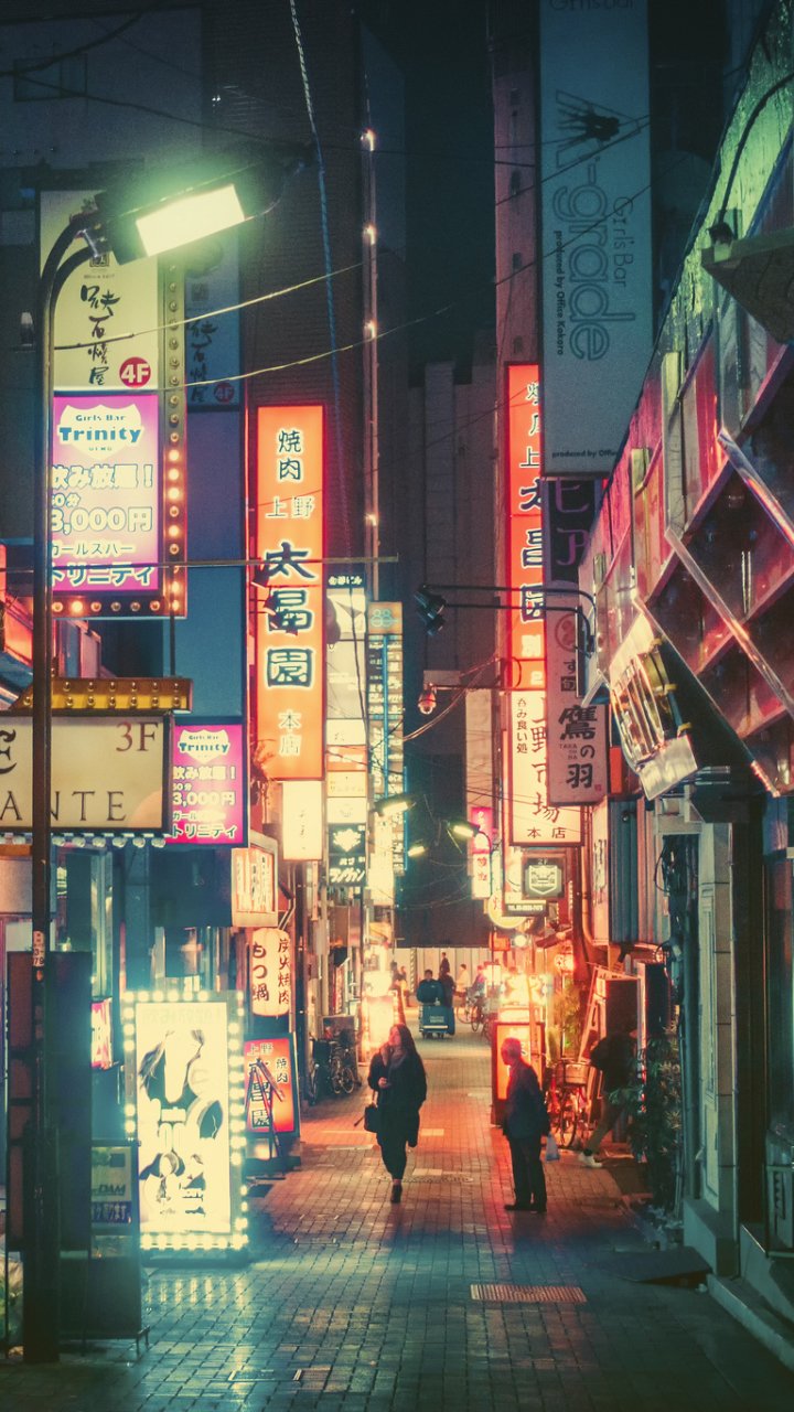 Neon-lit Tokyo street at night with vibrant signs and two people walking, captured in a vertical cityscape designed as a phone wallpaper for iPhones and Android devices.