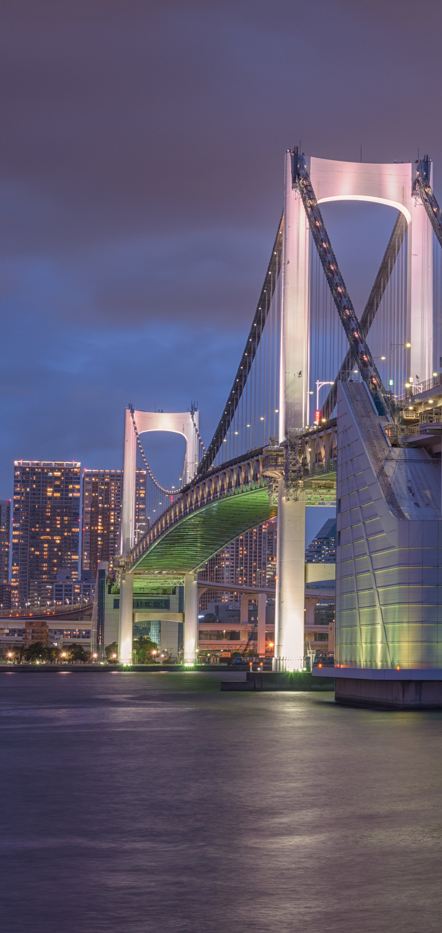 Night view of Tokyo's illuminated Rainbow Bridge spanning over calm waters with city lights in the background, designed as a phone wallpaper for iPhones and Android devices.