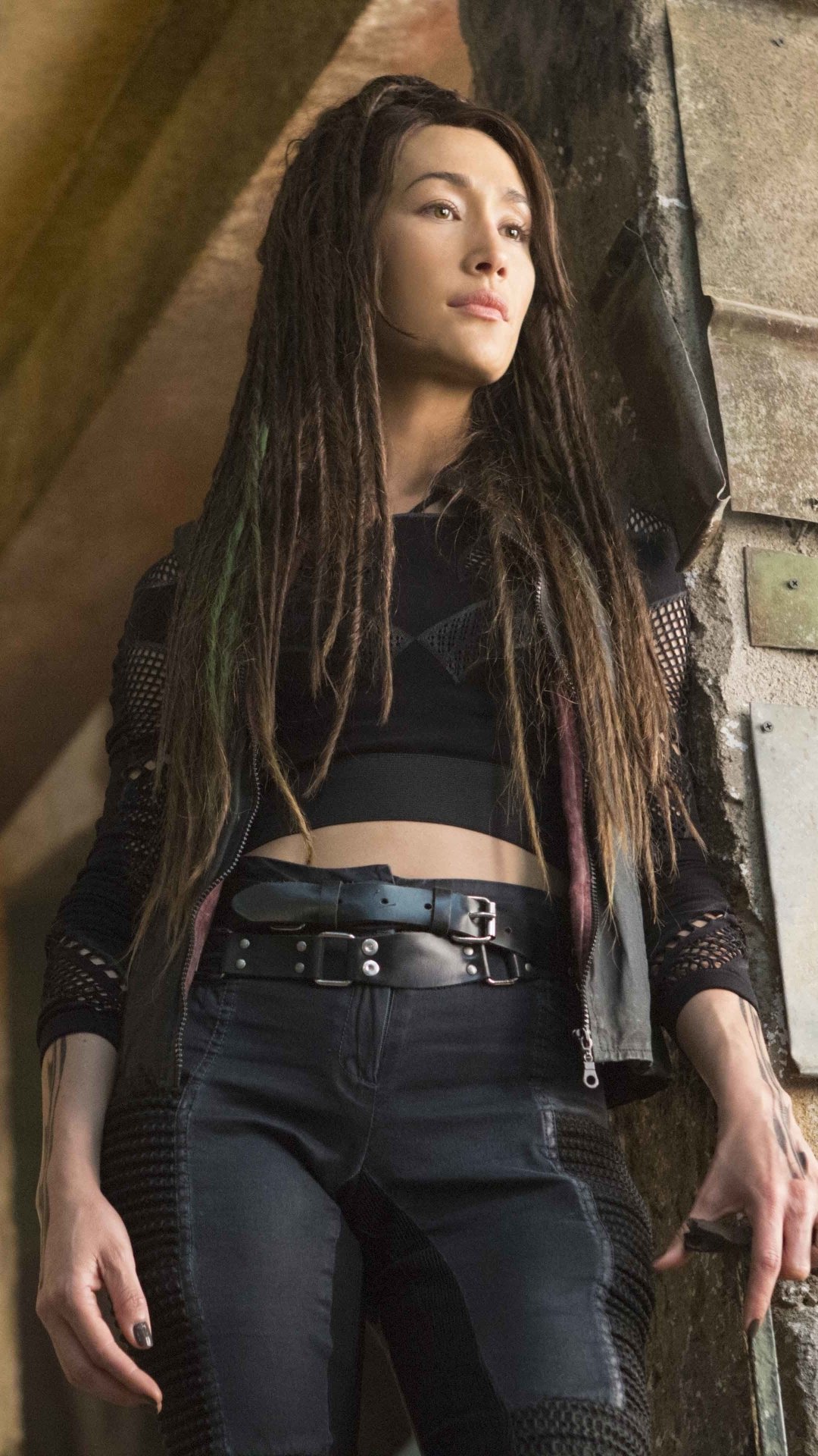 Phone wallpaper featuring Maggie Q as Tori from The Divergent Series: Allegiant, showcasing her in a dark outfit against a rustic stone background.