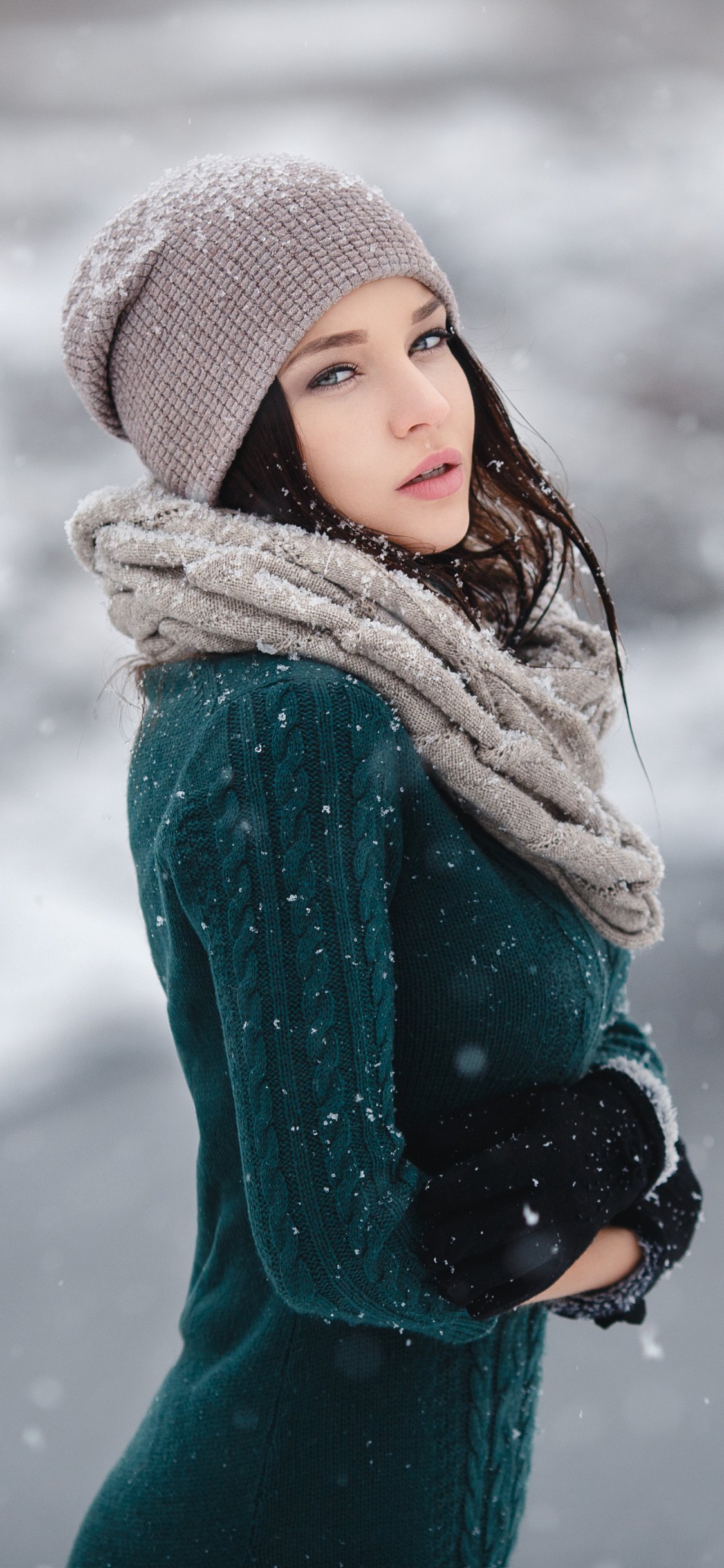 Phone wallpaper portrait of a woman model wearing a knit hat and chunky scarf, snowflakes drifting around her against a soft winter backdrop.