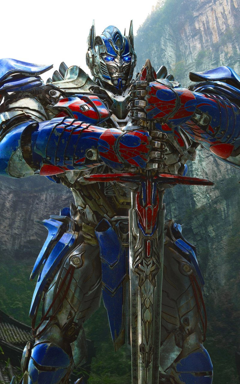 Optimus Prime from the Transformers movie, standing with a sword in a lush, mountainous background, designed as a phone wallpaper for iPhone and Android.