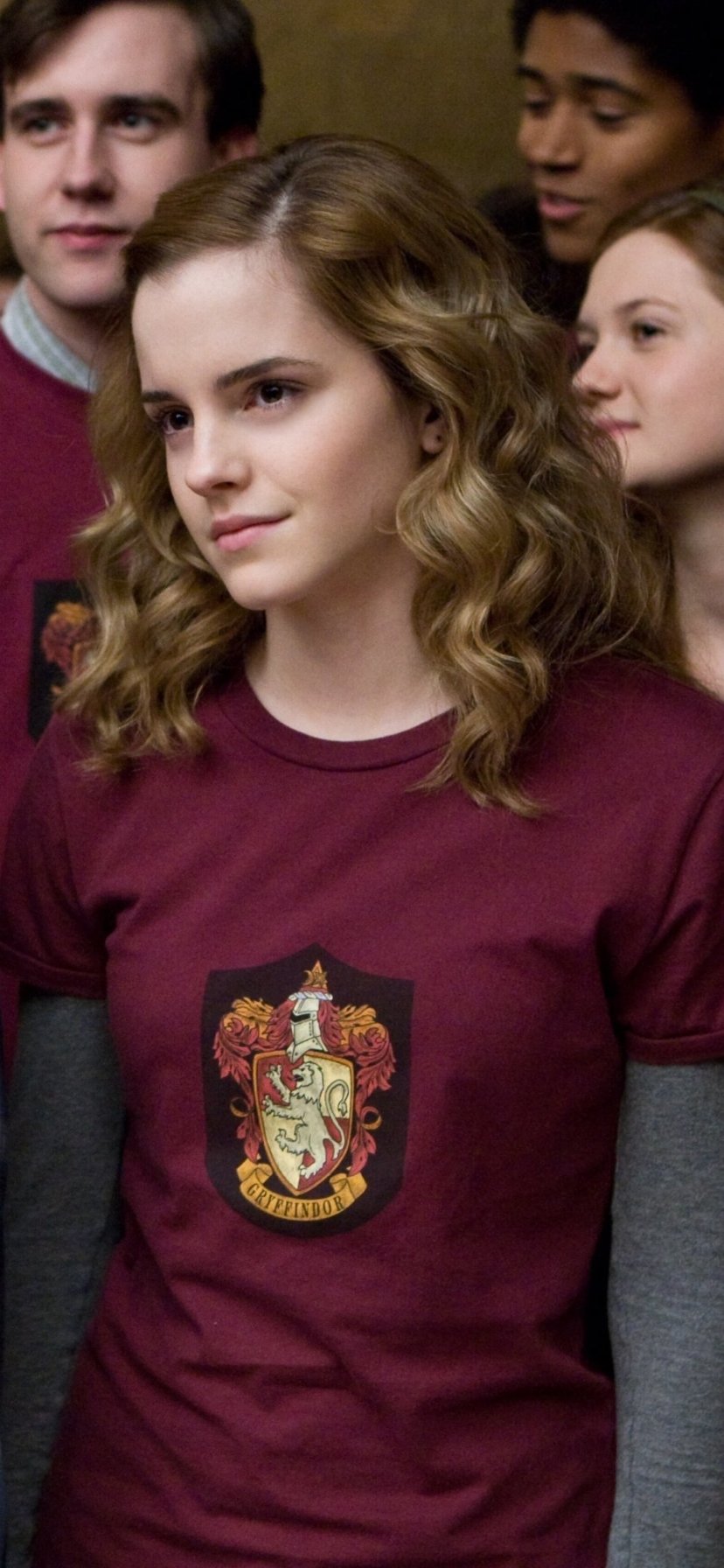 Phone wallpaper of Hermione Granger from the movie Harry Potter and the Half-Blood Prince: close portrait in a Gryffindor shirt with classmates blurred behind her.