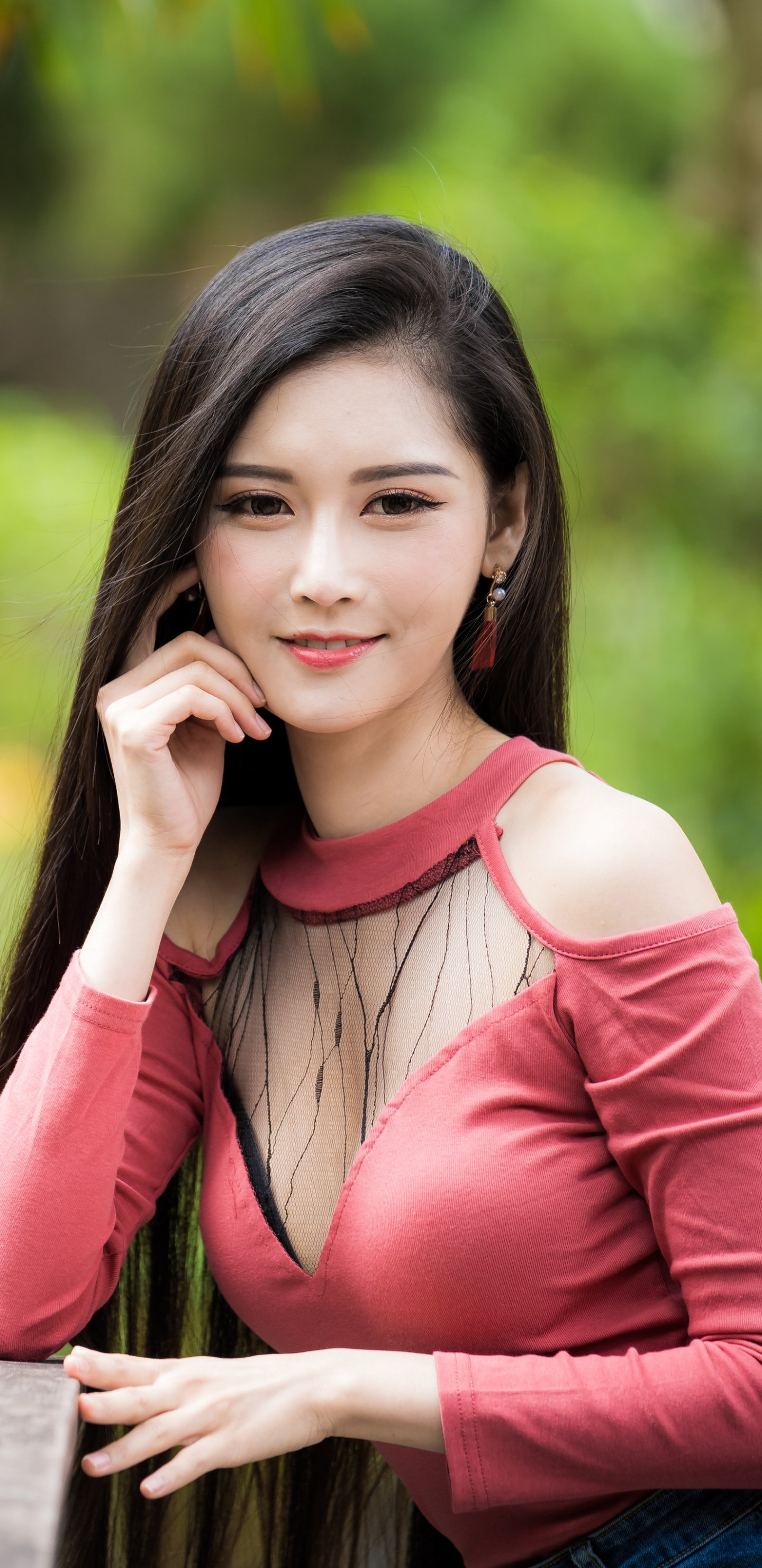 Phone wallpaper: portrait of a black-haired woman model smiling in a pink cold-shoulder top, hand to chin, with soft outdoor bokeh background.