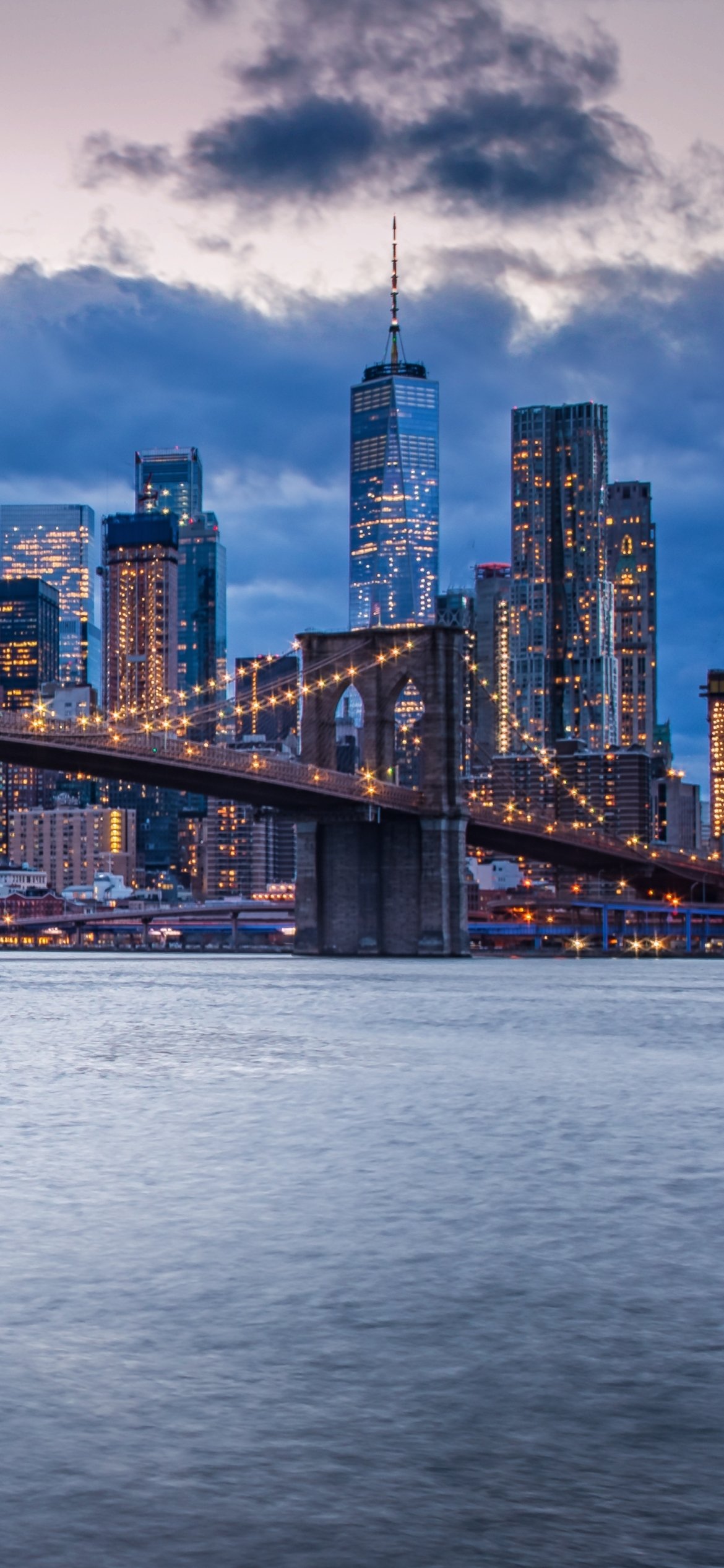 Phone wallpaper: New York City skyline at dusk — the man-made Brooklyn Bridge spans the East River with illuminated skyscrapers, including One World Trade Center, rising behind.
