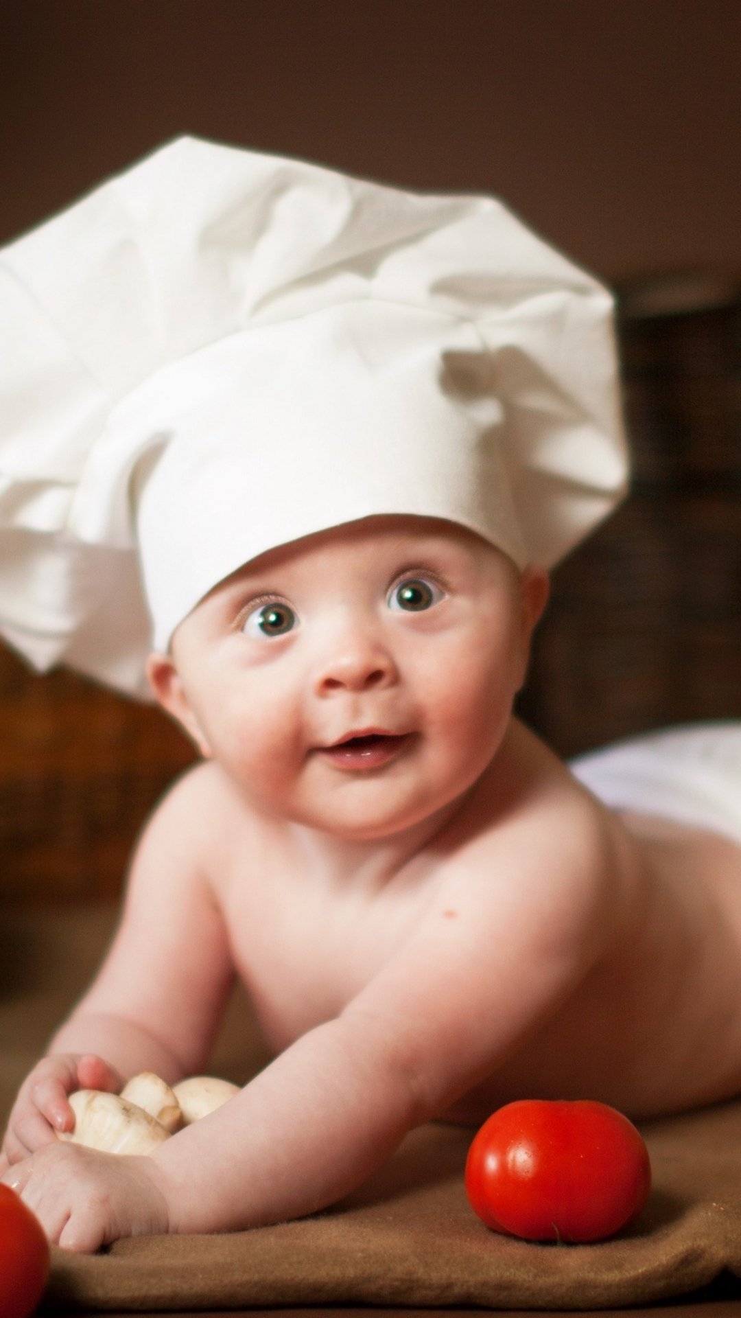 Baby photography: a wide-eyed infant in a chef's hat propped on elbows with tomatoes nearby, warm playful phone wallpaper.