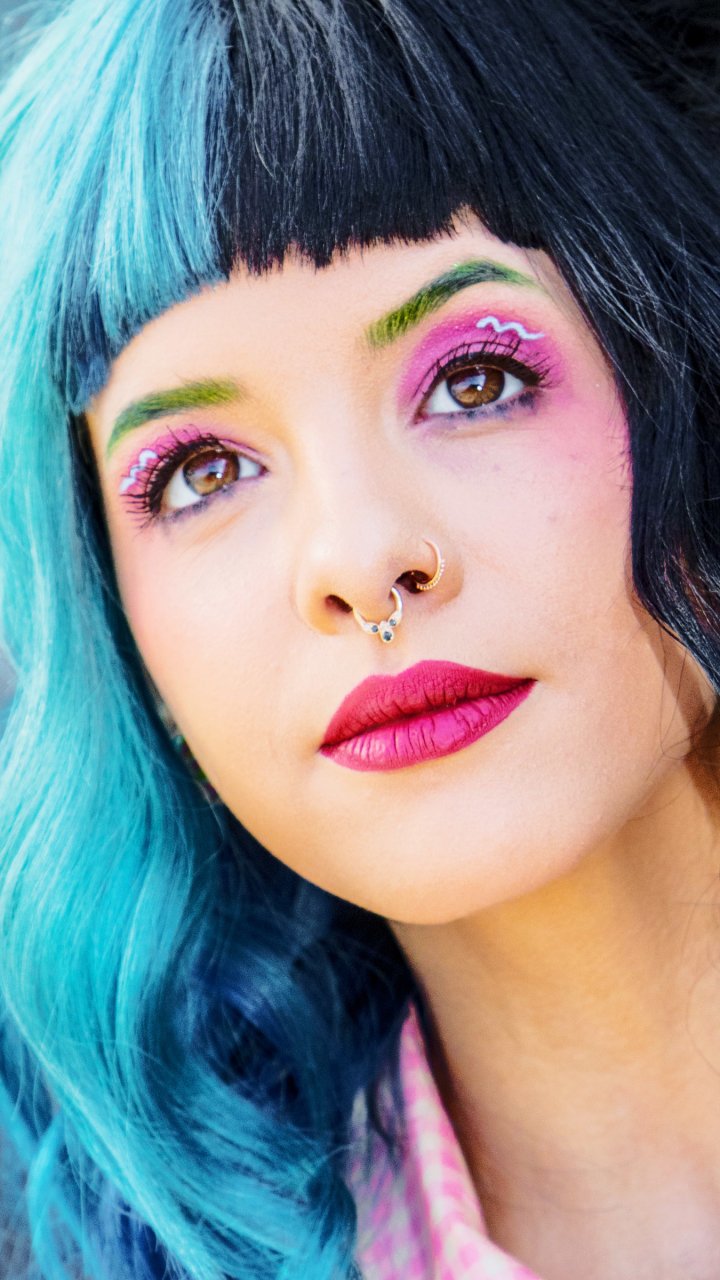 Phone wallpaper close-up of a face with half teal, half black hair, bright pink lipstick, green and purple eye makeup and a septum ring — polished pop-singer–inspired look.