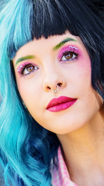 Phone wallpaper close-up of a face with half teal, half black hair, bright pink lipstick, green and purple eye makeup and a septum ring — polished pop-singer–inspired look.