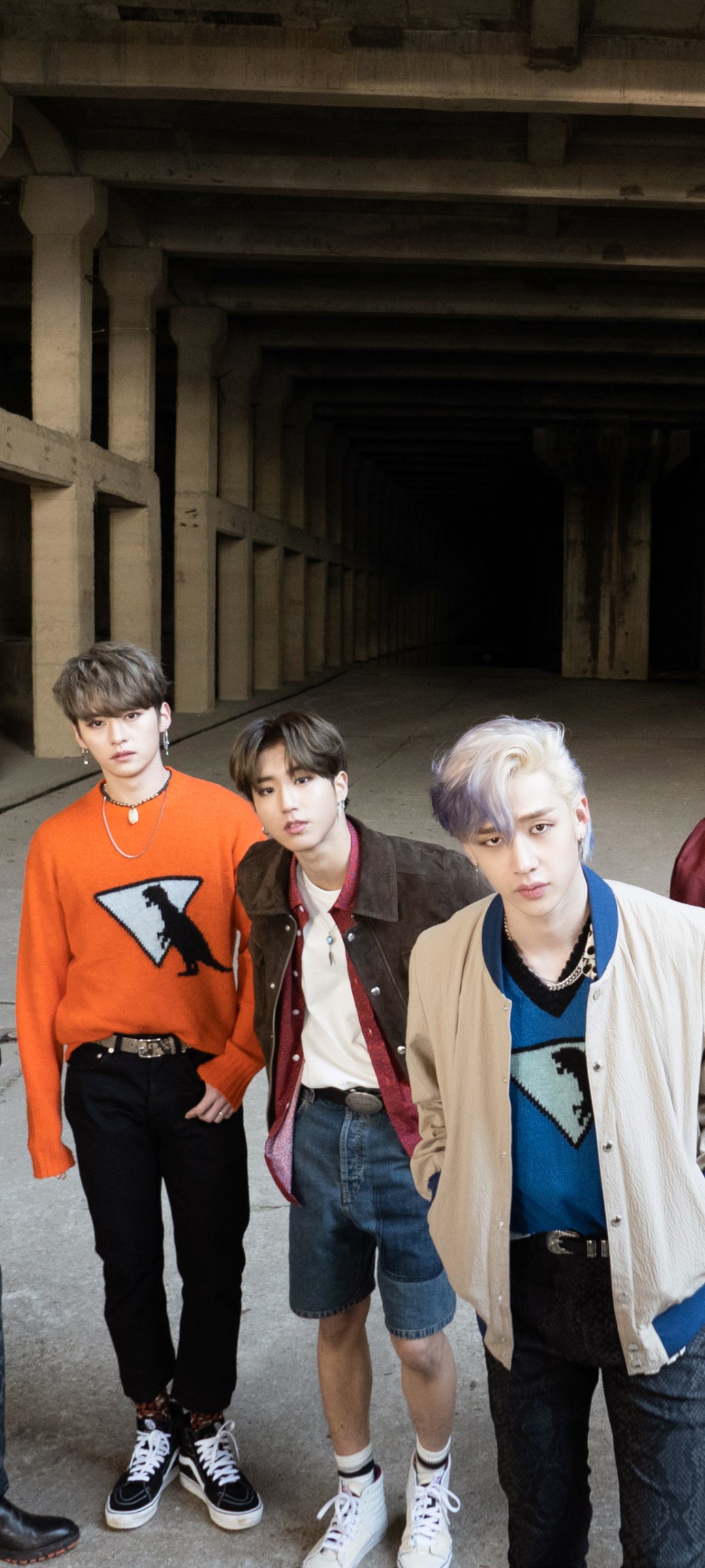Phone wallpaper of a K-pop boy band: four stylish young men in casual streetwear posing in a dim, concrete industrial tunnel with a moody music vibe.