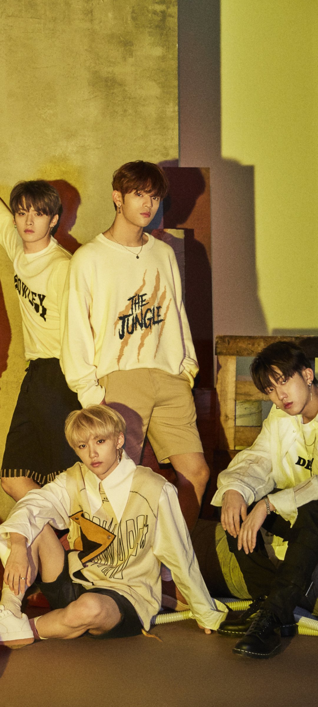 Phone wallpaper of a K-pop boy band in coordinated streetwear, four members posed against warm yellow-green lighting, evoking a moody, music-focused promotional vibe.