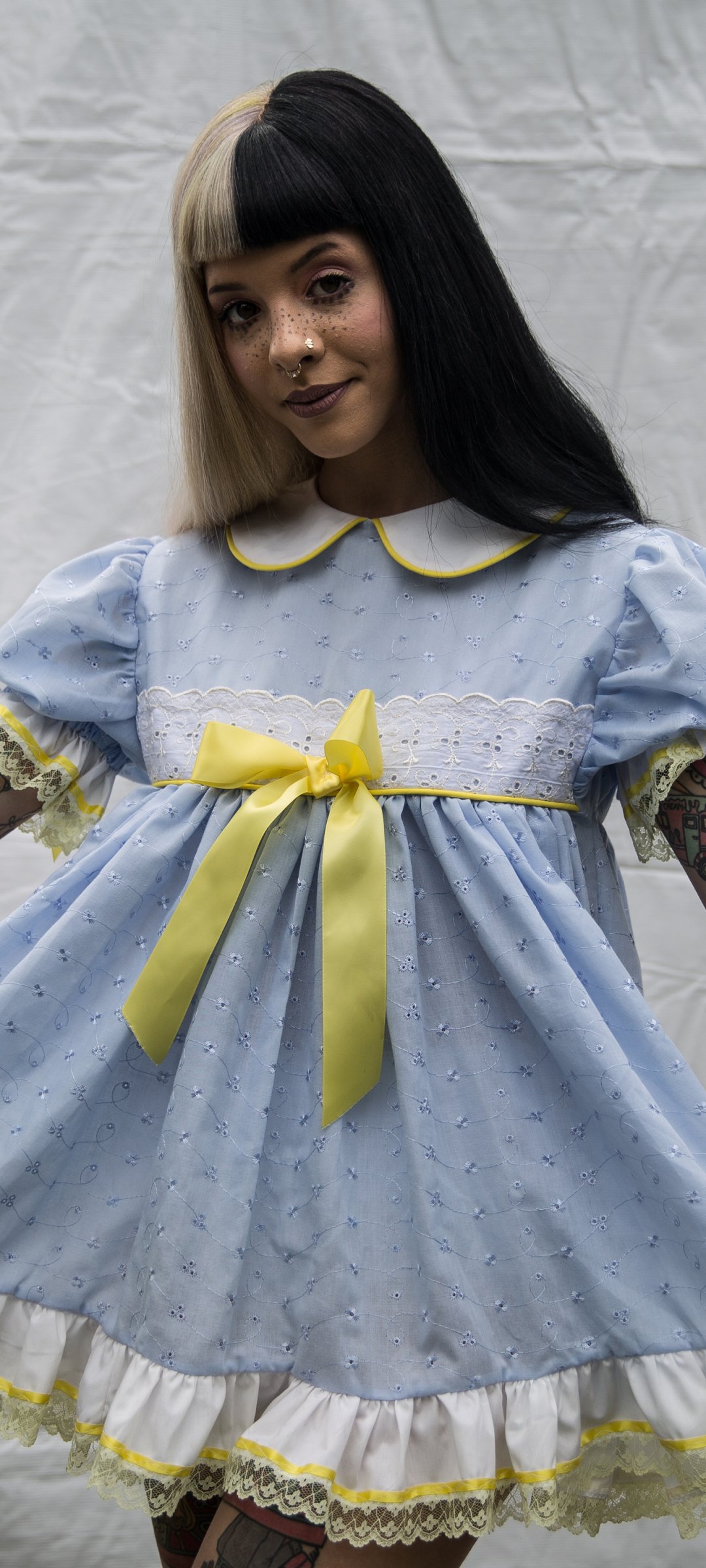 Phone wallpaper of an American singer in a two-tone wig wearing a blue ruffled dress with yellow bow, visible arm tattoos — stylized music portrait.