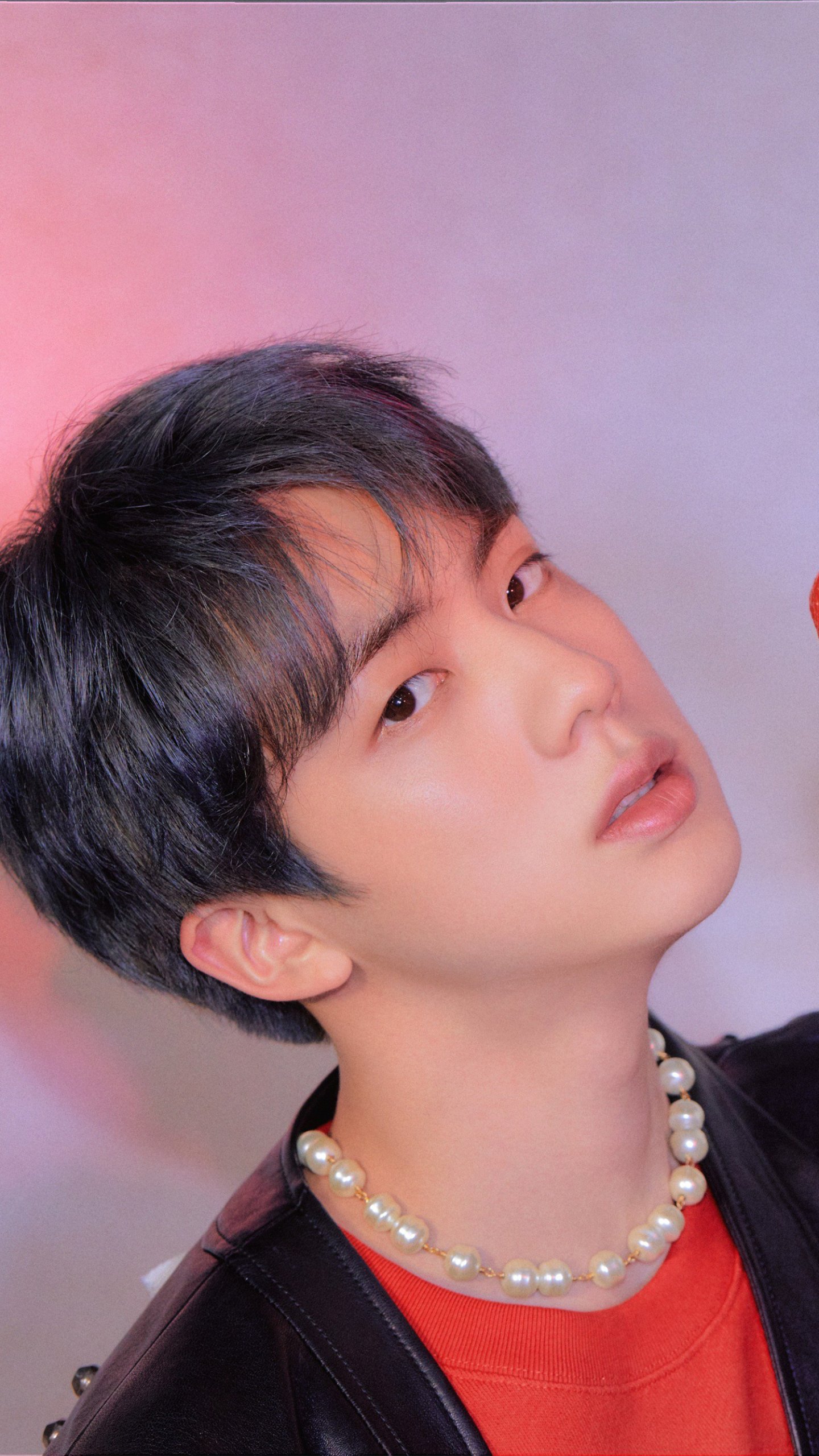 Phone wallpaper portrait of a K-pop singer gazing over his shoulder against a soft pastel backdrop, wearing a pearl necklace and leather jacket.