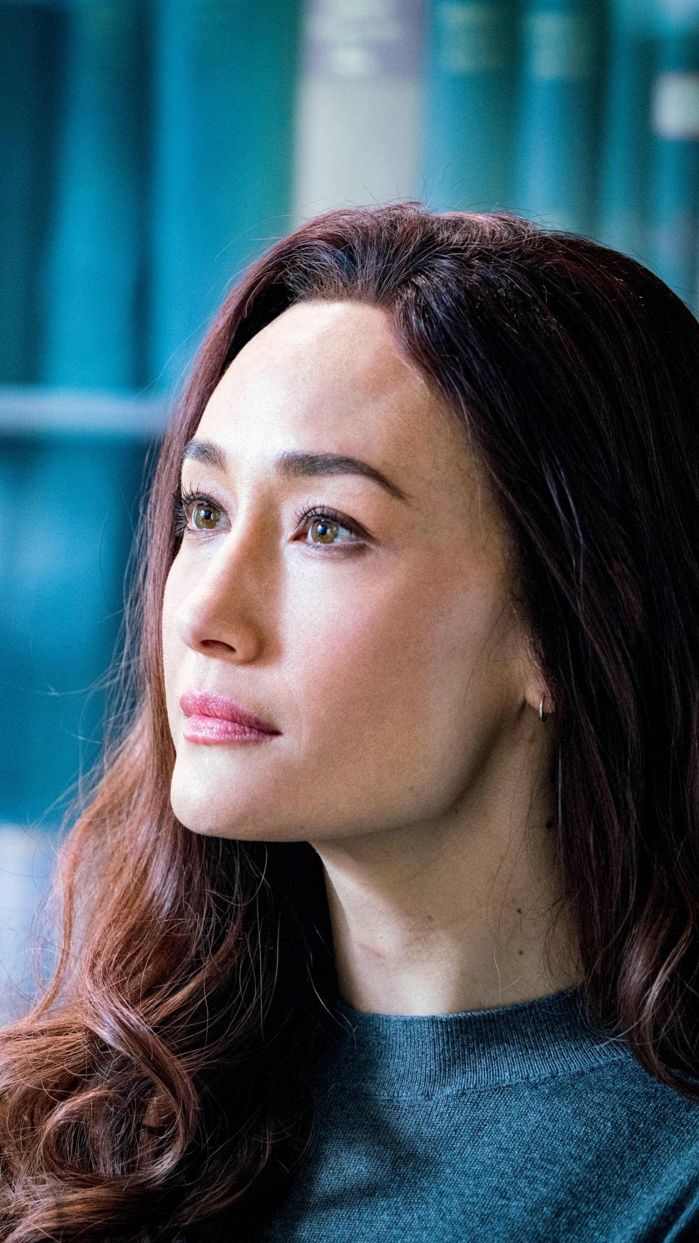 Phone wallpaper featuring Maggie Q from the movie The Protégé, with a close-up of her face against a softly blurred blue background.